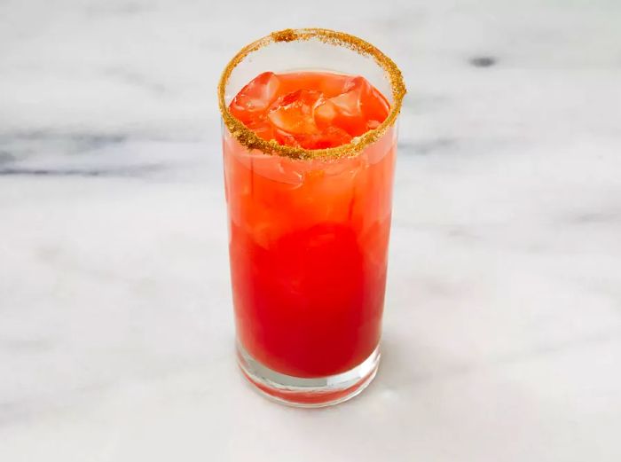 An overhead shot of tomato-clam juice being poured into a spiced highball glass containing vodka and Worcestershire sauce.