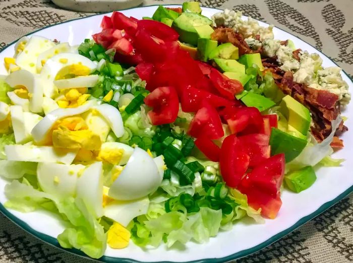 a beautifully arranged salad with hard-boiled egg, tomatoes, chicken, avocado, crumbled bacon, and blue cheese