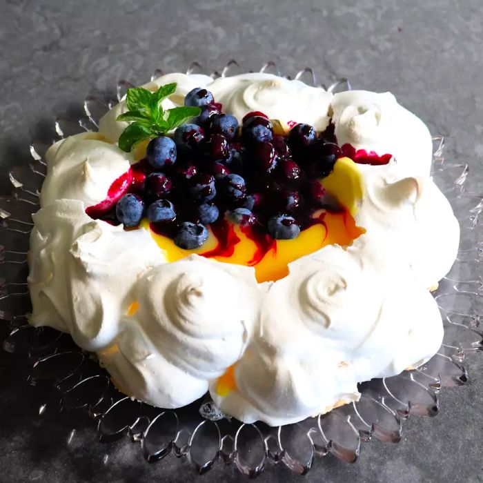 This Lemon Blueberry Pavlova features a delicate meringue shell filled with a tangy lemon curd, fresh blueberries, and a drizzle of blueberry sauce, all garnished with a sprig of fresh mint for an extra touch of elegance.