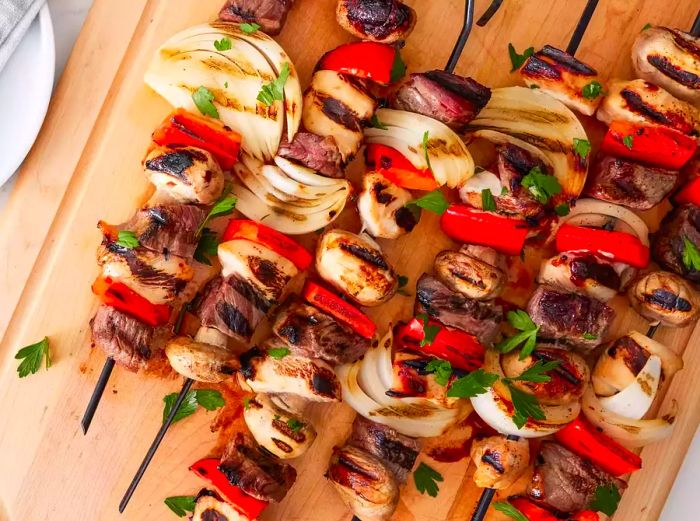 A cutting board with grilled chicken, beef, and vegetable kabobs ready to serve