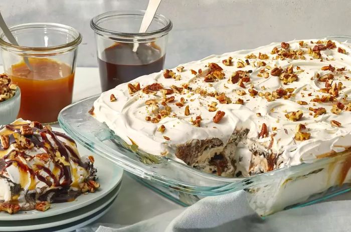 A side view of an ice cream sandwich cake in a clear 9x13 casserole dish, with a slice plated on the side, topped with chocolate and caramel sauce.