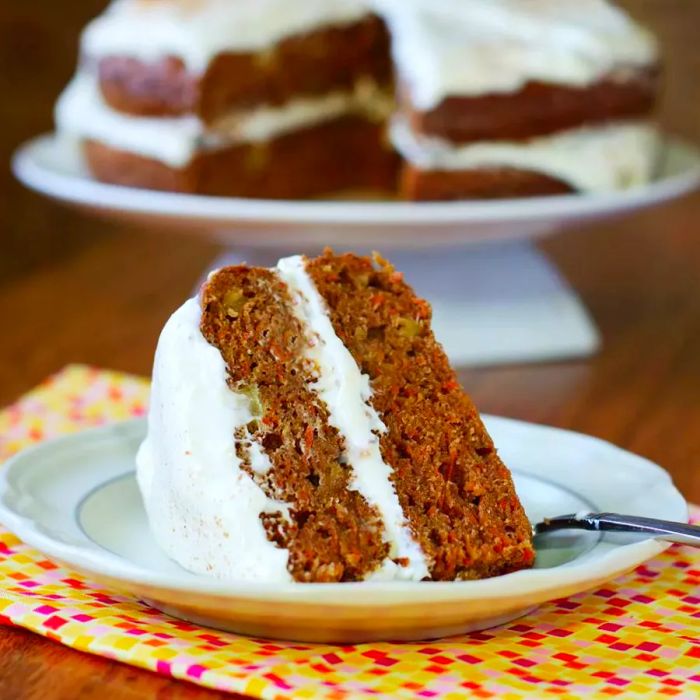 A slice of carrot cake served on a white plate with the whole cake in the background