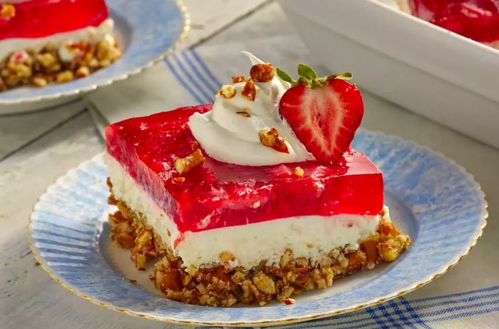 A close-up of a slice of Judy's strawberry pretzel salad, topped with whipped cream, fresh strawberries, and crunchy pretzel pieces.