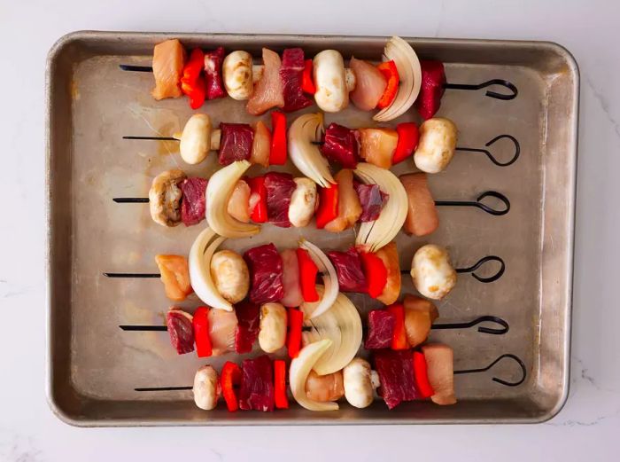 A baking sheet with raw beef, chicken, and vegetable kabobs ready for grilling