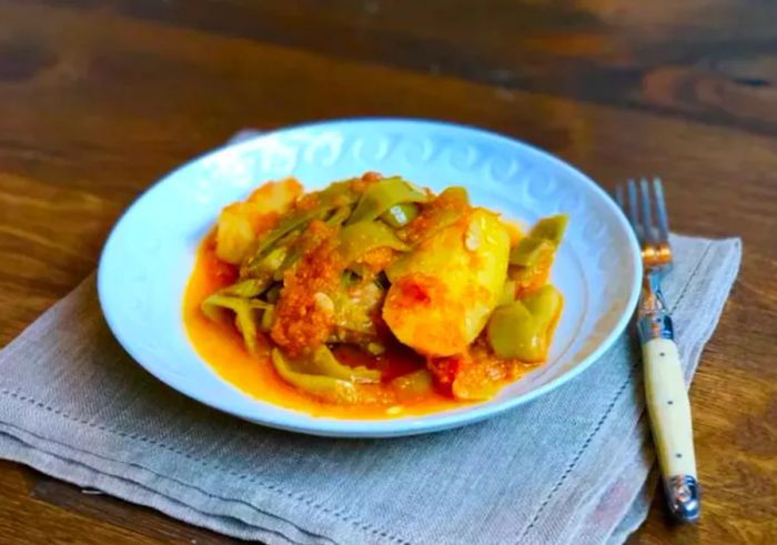 A white bowl filled with crispy potato wedges and green beans simmered in a savory tomato sauce