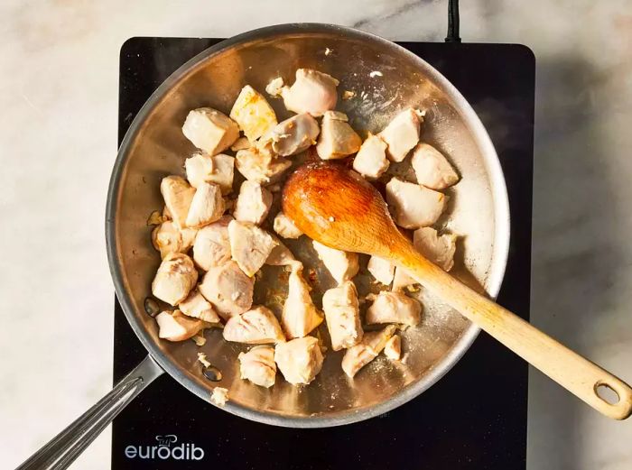 A pan of seared chicken cubes being stirred with a wooden spoon