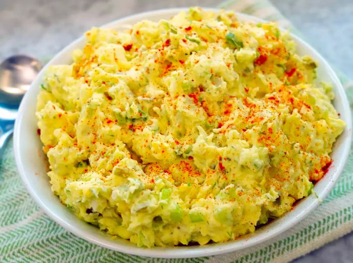 A bowl of smooth, creamy potato salad topped with a sprinkle of paprika, served in a white dish with a serving spoon on the side