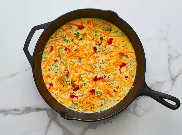 A top-down view of the egg mixture in a cast iron skillet, topped with cheese, bacon, and green onions.