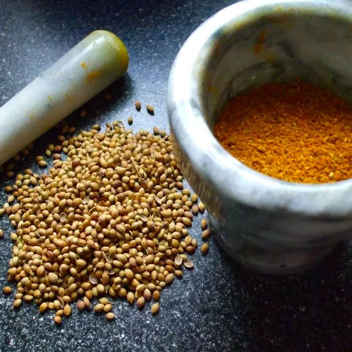 A white marble mortar and pestle with golden powder and coriander seeds resting beside it