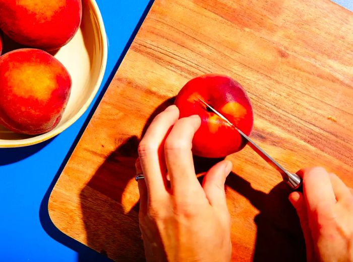 Holding a peach and a paring knife, ready to slice from the stem to the blossom end.