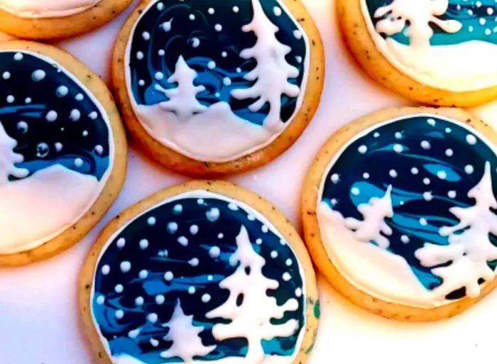 Sugar cookies decorated with winter landscapes: a serene night sky filled with falling snowflakes and pine trees blanketed in snow.