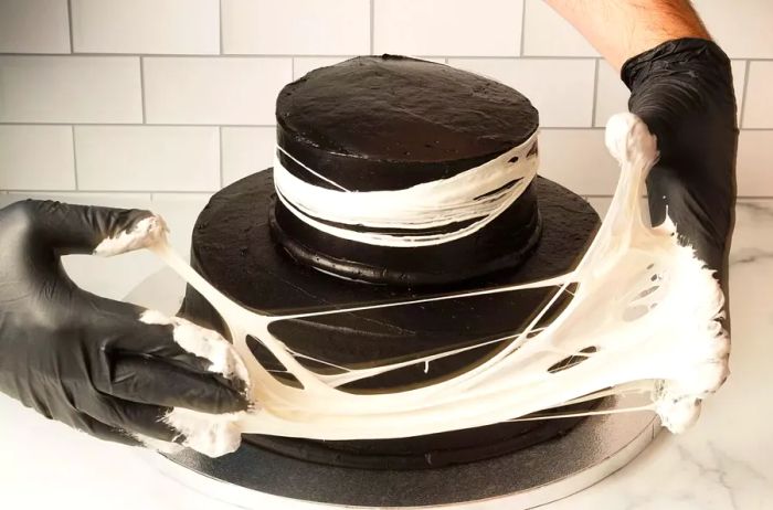 Using gloved hands to stretch the melted marshmallow into a spiderweb decoration on a two-tier dark chocolate cake for Halloween