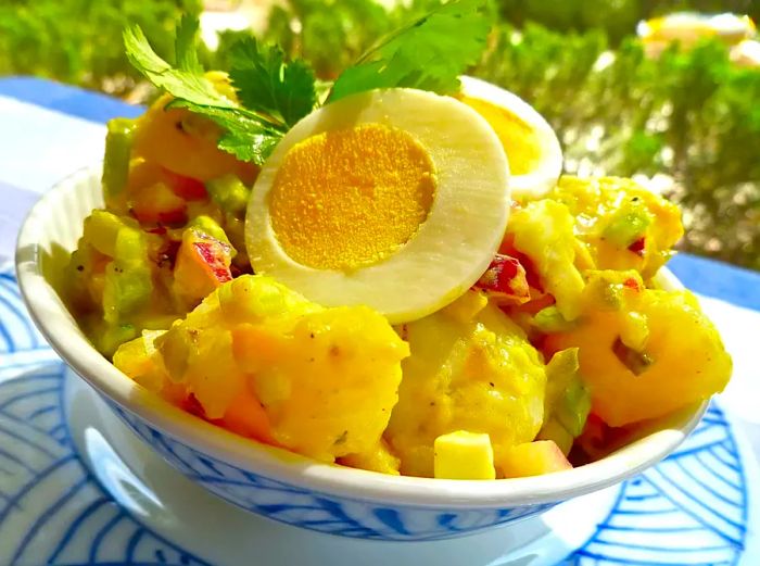 A serving of classic Old-Fashioned Potato Salad topped with slices of hard-boiled egg and a sprig of fresh parsley, served in a blue-and-white bowl