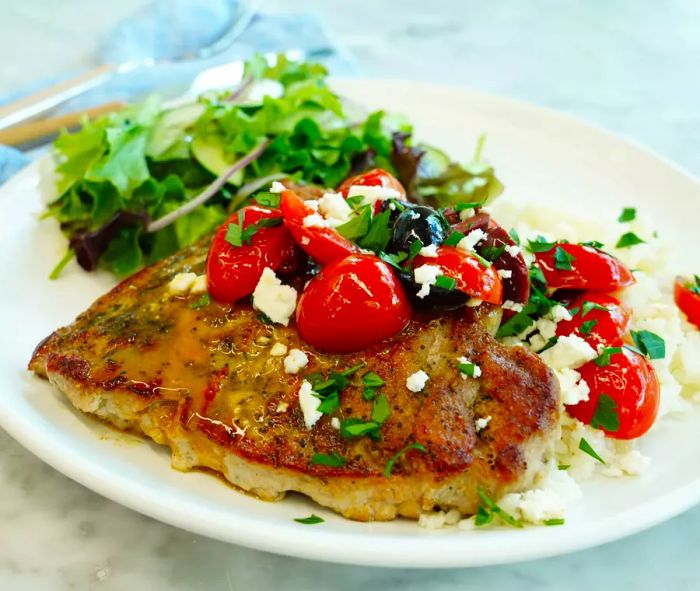 A golden-brown pork chop garnished with halved cherry tomatoes, crumbled feta cheese, and kalamata olives, served with a side of mixed greens