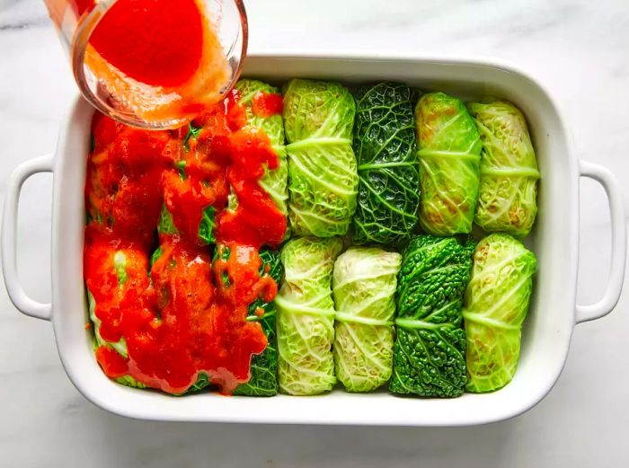 A high-angle shot of stuffed cabbage rolls in a baking dish as tomato sauce is poured over them.