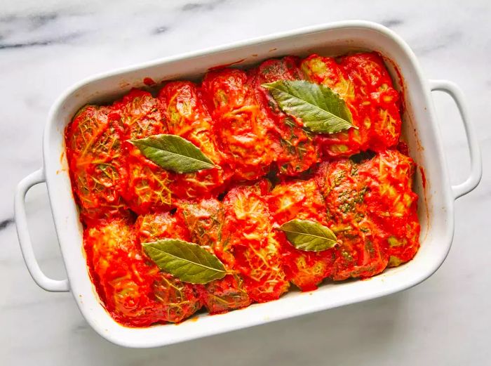 A top-down view of a baked dish of cabbage rolls, smothered in tomato sauce with bay leaves.