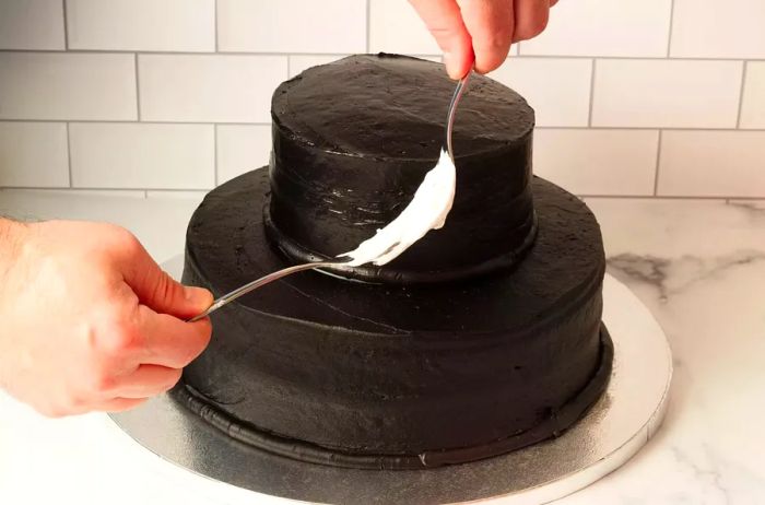 Using two forks to spread melted marshmallow over a two-tier dark chocolate cake to create a spiderweb decoration