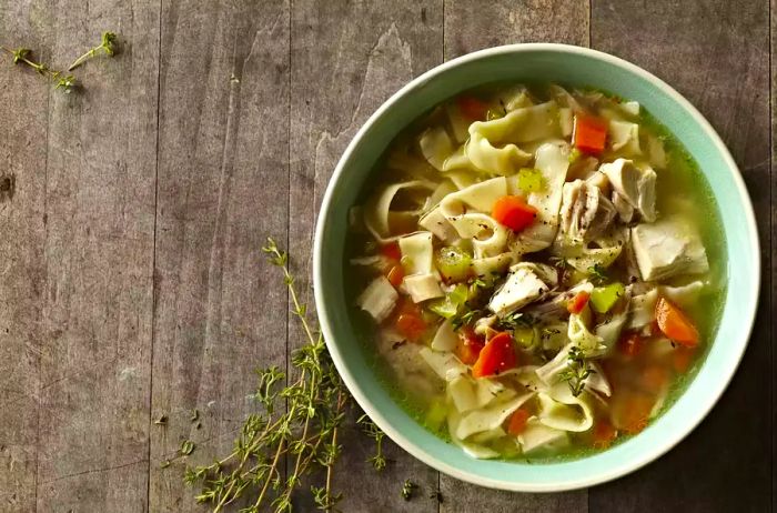 A bowl of homemade chicken soup, filled with noodles, carrots, celery, and fresh thyme