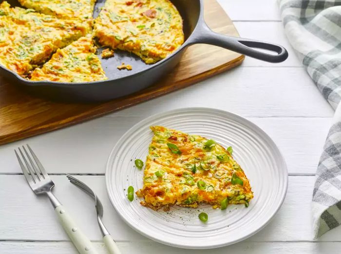 A slice of bacon cheese frittata served on a plate, with the rest remaining in the cast iron skillet behind it.