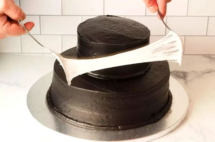 Using two forks to stretch melted marshmallows and form a spiderweb decoration on a two-tier dark chocolate cake for Halloween