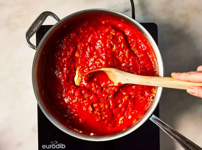 A wooden spoon mixing tomato sauce into a pan of spiced onions