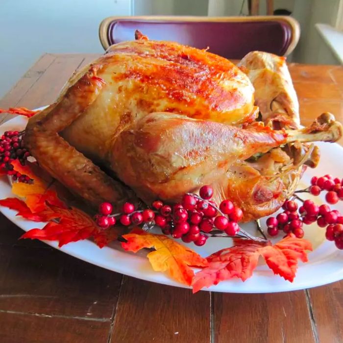 A beautifully golden roast turkey, served on a white platter with festive fall leaves and cranberry garnishes.