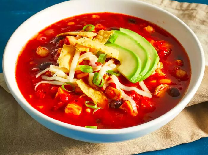 A close-up, low-angle shot of a bowl of chicken tortilla soup topped with shredded cheese, crispy tortilla strips, and avocado slices.