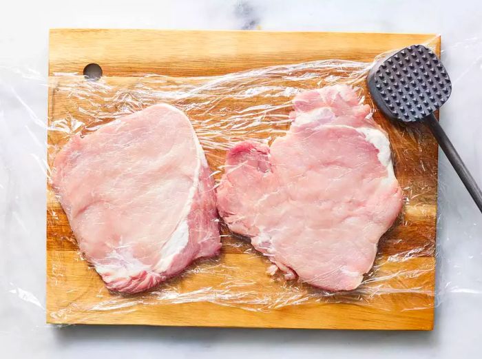 Pounding chicken cutlets thin on a cutting board.