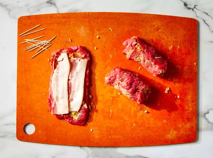 A top-down view of a steak with bacon, and two steaks rolled up and held together with toothpicks on a cutting board