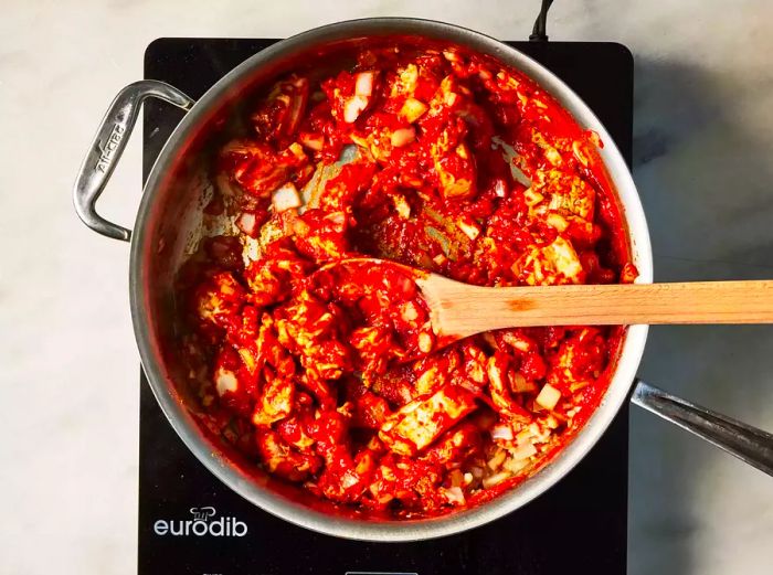 A pan filled with chicken pieces, garlic, and onions simmering in a rich blend of tomato paste and aromatic spices.