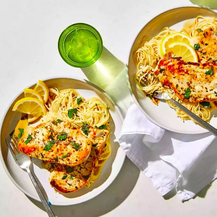 Two white plates featuring pan-fried chicken breasts alongside spaghetti noodles, decorated with lemon slices and parsley.