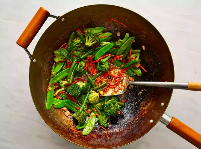 A top-down view of vegetables being mixed with soy sauce and stir-fried in a wok.