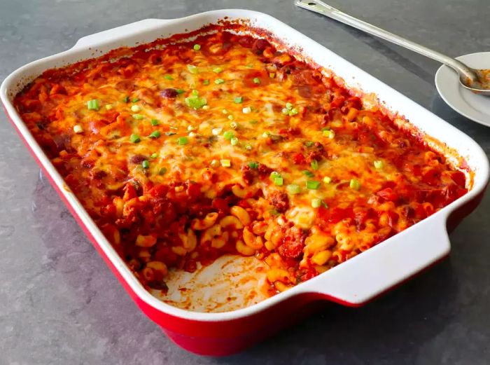 A red rectangular casserole dish filled with chili dog mac and cheese, with one serving already removed.