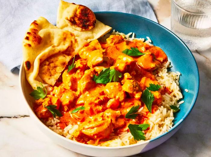 A bowl of slow-cooked butter chicken served over rice, accompanied by naan bread.