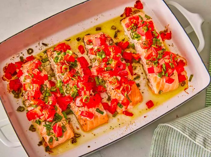 Bird's-eye view of four salmon fillets in a baking dish, topped with diced tomatoes and green onions.