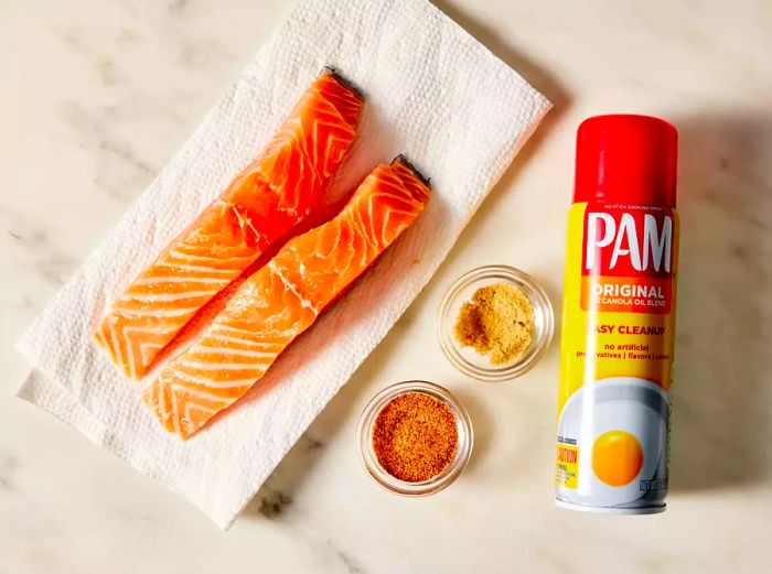 A top-down view showcasing salmon fillets, seasonings, and cooking spray on a marble countertop.