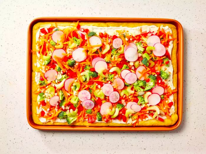 A jelly roll pan featuring a veggie pizza topped with freshly chopped broccoli, radishes, onions, bell peppers, carrots, and celery.