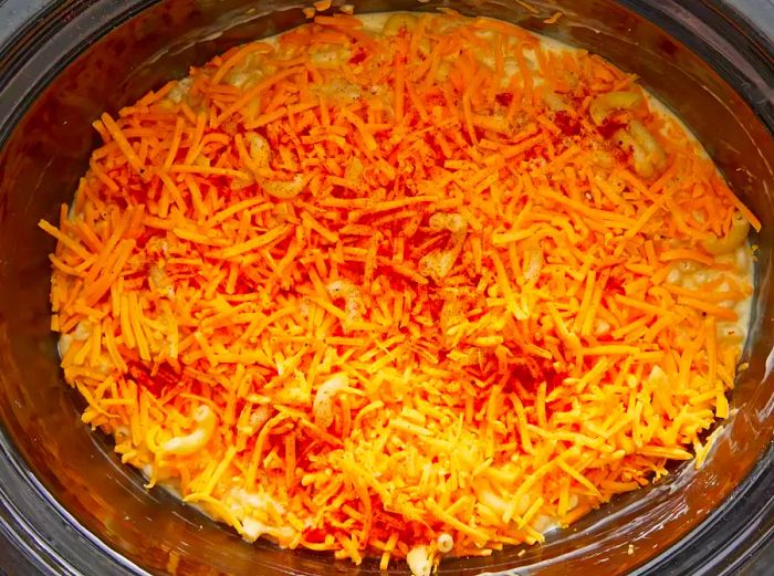 A top-down view of shredded cheddar cheese sprinkled over a crockpot filled with creamy macaroni and cheese sauce.