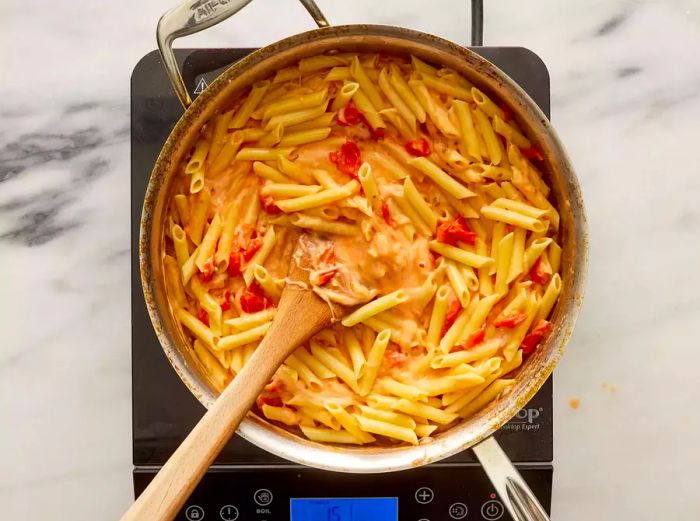 A pan with penne pasta being combined with blistered grape tomatoes and melted cheese