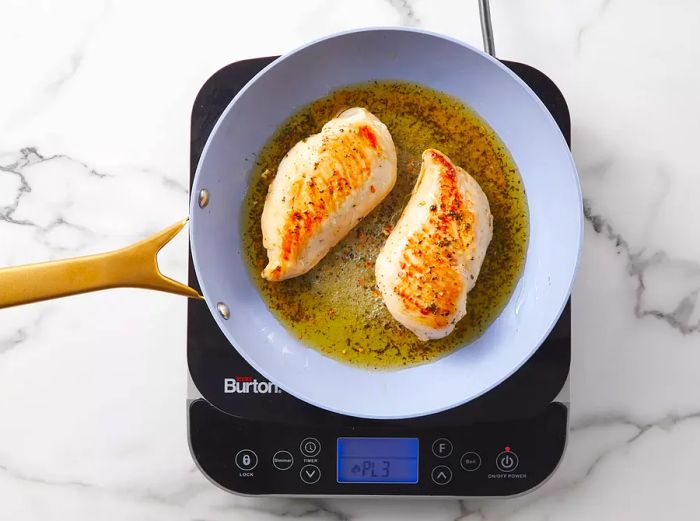 Two golden-brown chicken breasts cooking in a hot skillet.