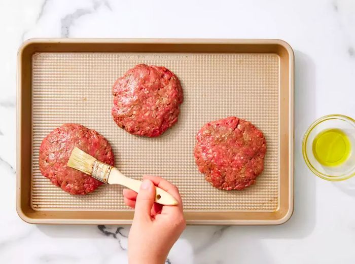 Brushing oil onto uncooked hamburger patties on a baking tray