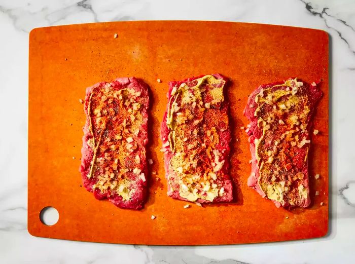 A top-down view of steak pieces coated with mustard, onion, paprika, salt, and pepper on a cutting board