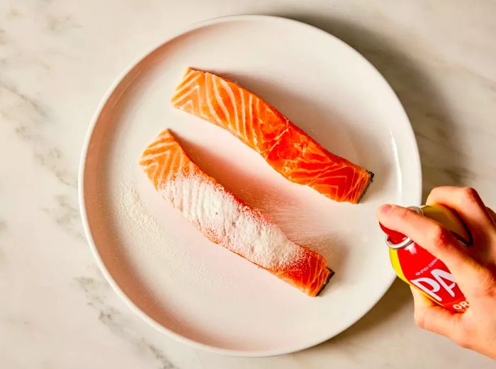 Two salmon fillets on a white plate being misted with cooking spray.