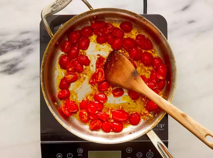 A pan with grape tomatoes, garlic, and oil being stirred with a wooden spoon