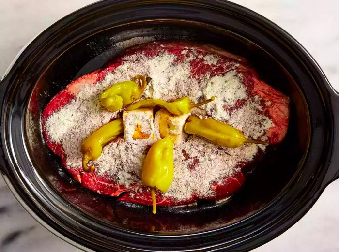 A slow cooker with a roast topped with butter, pepperoncini peppers, ranch seasoning, and au jus mix.