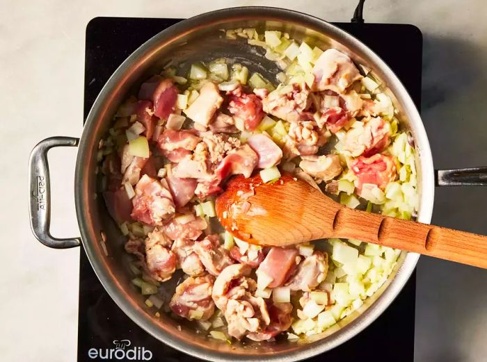 A large skillet containing chicken pieces, onion, and garlic being stirred with a wooden spoon.