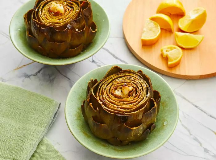 Two roasted artichokes served in small bowls, with a lemon on a cutting board beside them.