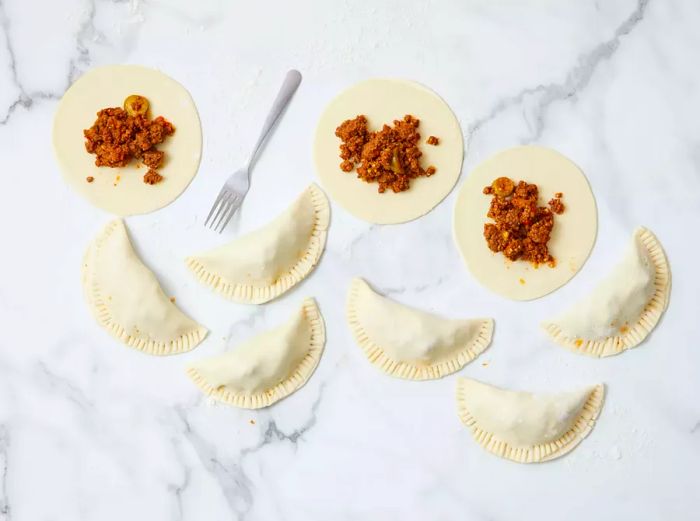 A partially opened empanada filled with seasoned beef, surrounded by folded and sealed raw empanadas