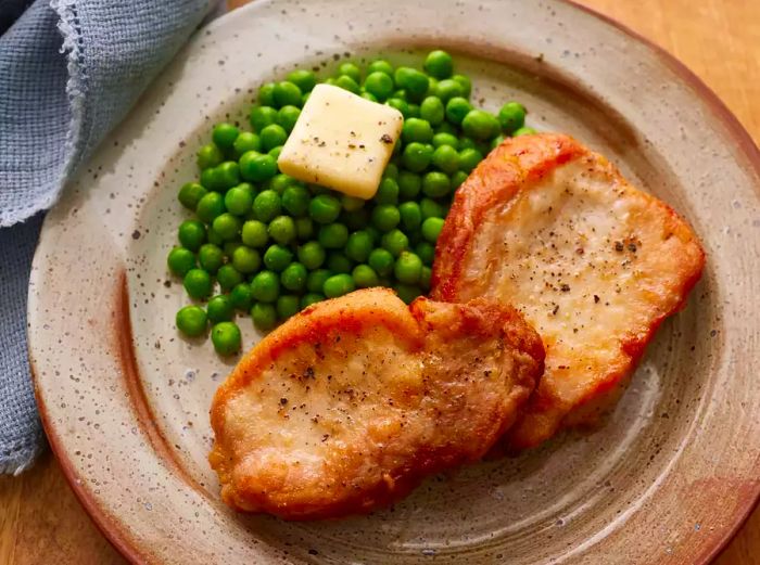 A close-up of a plate with golden-brown fried pork chops, served with green peas and a dollop of butter.