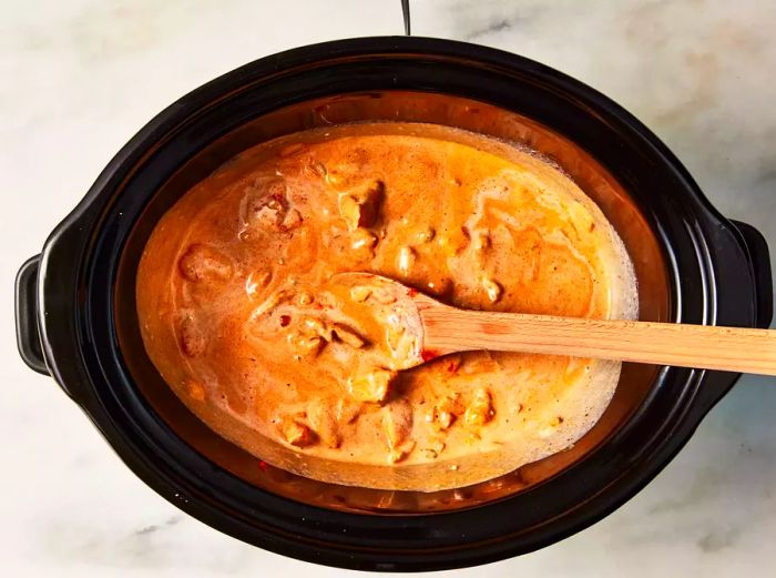 A crockpot with slow cooker butter chicken being stirred with a wooden spoon.